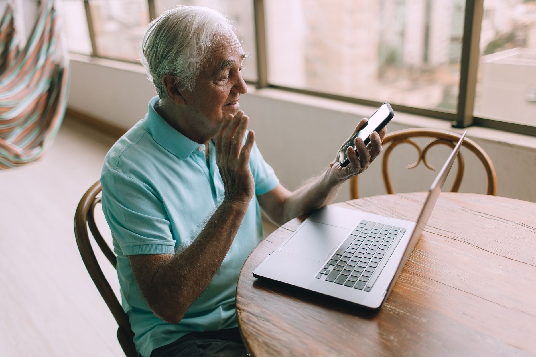 Senior mężczyzna rozmawia na wideo przez smartfon i laptop w domu